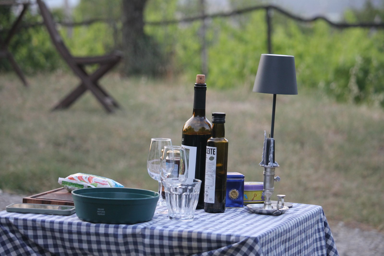 Table with wine, olive oil, glasses and a lamp in front of a vineyard